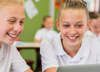 Two female students sat in a classroom, with other students visible in the background, looking at a laptop screen and smiling