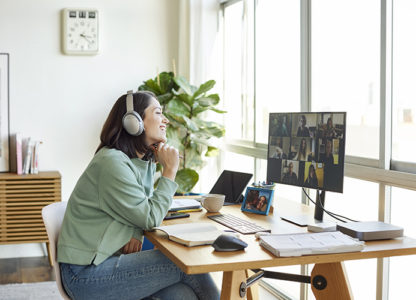 Smiling businesswoman discussing with colleagues through video call. Female professional is sitting in bright home office. She is listening through headphones.